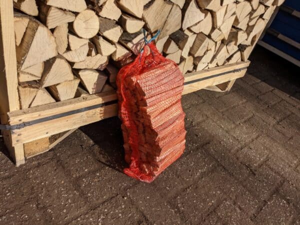 Een stapel gekapt brandhout ligt netjes op een houten pallet. Vooraan leunt een rode netzak met het label "Aanmaakhout 3kg - Zakje Aanmaakhout - Snel Vuur Aansteken" tegen de stapel op een zonovergoten geplaveid oppervlak, klaar voor snelle ontsteking.